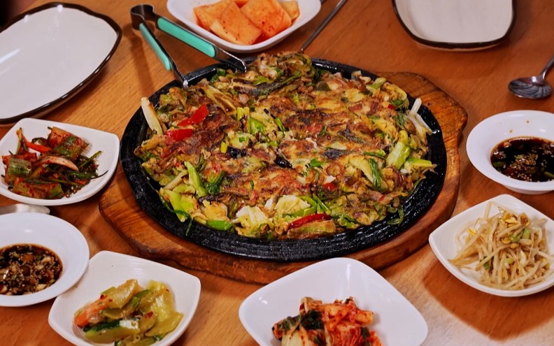 A fried pancake-like dish on a plate surrounded by vegetables and sauces.
