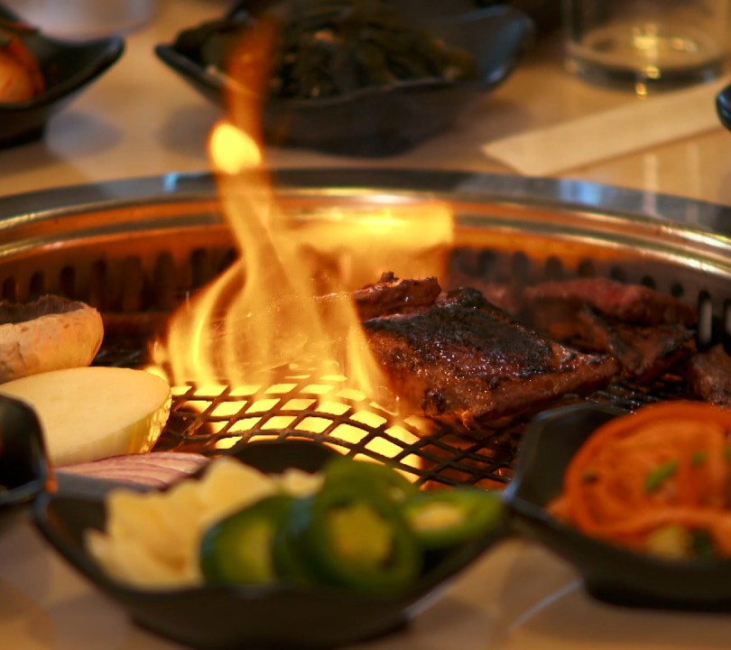 A piece of meat being cooked by flame from a grill, located in the middle of a table.